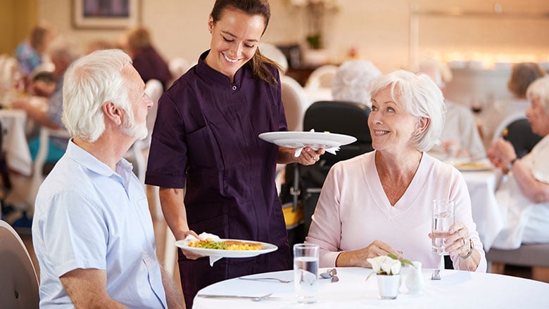 Senior Couple Being Served With Meal By Carer In Dining Room Of Retirement Home