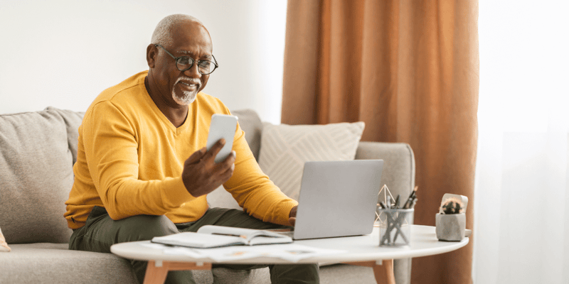Older man using a smartphone and laptop while seated on a sofa